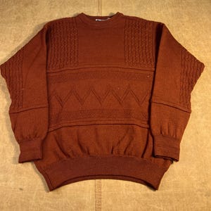 May include: A rust-colored sweater with textured knit panels on the shoulders and sleeves. The sweater features a horizontal band and a zig-zag pattern across the chest. The cuffs and hem are ribbed.