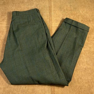May include: Green plaid trousers with a classic fit. The pants feature a subtle pattern in shades of green and blue. The trousers are folded, showcasing the tailored design and the fabric's texture. A timeless piece for a polished look.