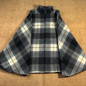 May include: A gray, black, and cream plaid cape with a high collar and a zipper. The cape has a wide, flowing design with a classic plaid pattern. The fabric appears to be a soft, woven material.