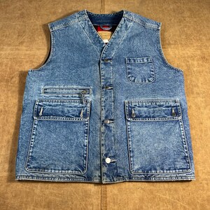 Könnte beinhalten: Eine blaue Jeansweste mit mehreren Taschen und einer Knopfleiste. Die Weste hat einen V-Ausschnitt und eine Reißverschlusstasche auf der linken Seite. Die Weste hat einen verblichenen, abgenutzten Look.
