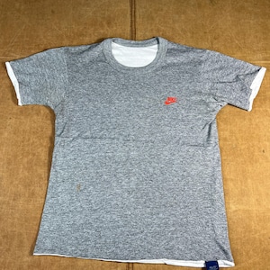 May include: Gray t-shirt with white trim on the collar and sleeves. A small red Nike logo is on the left chest. A blue tag with white text is attached to the bottom hem.