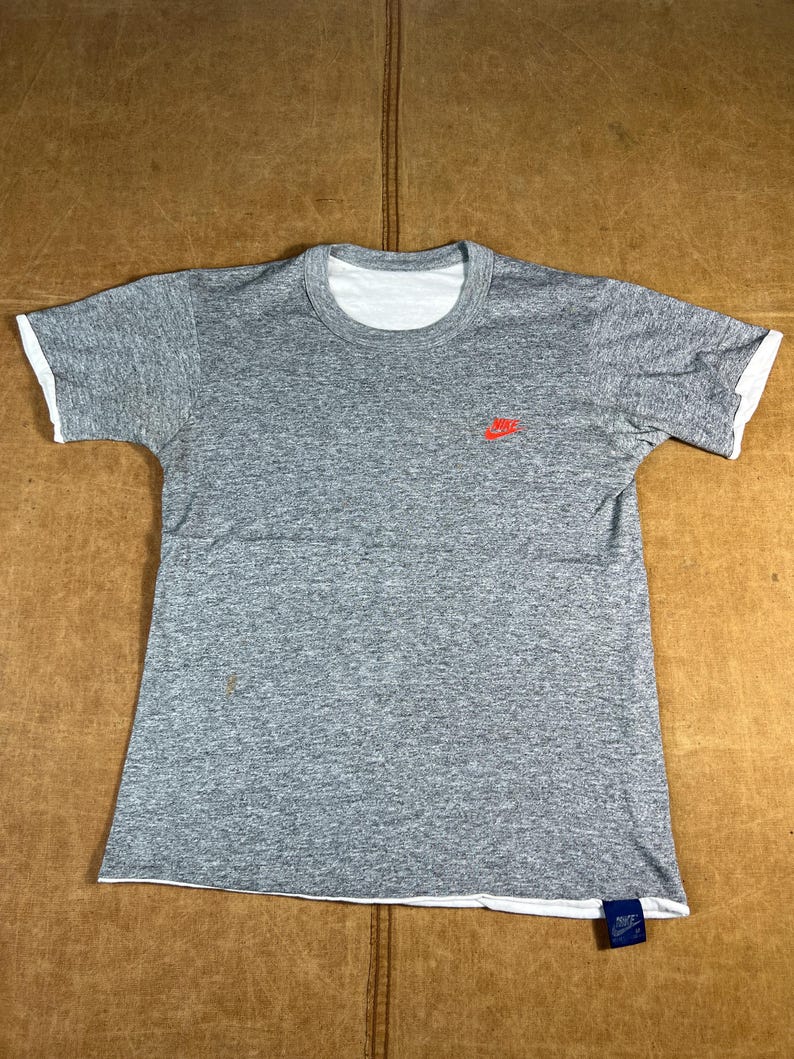 May include: Gray t-shirt with white trim on the collar and sleeves. A small red Nike logo is on the left chest. A blue tag with white text is attached to the bottom hem.