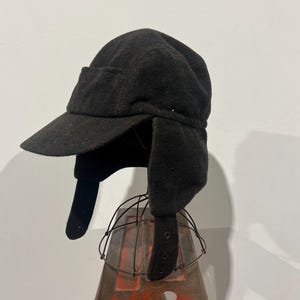Peut inclure: Une casquette d'hiver gris anthracite foncé dotée d'une visière rigide à l'avant et de deux rabats latéraux qui pendent vers le bas. La casquette est fabriquée dans une matière texturée semblable à du feutre et comporte des œillets métalliques.