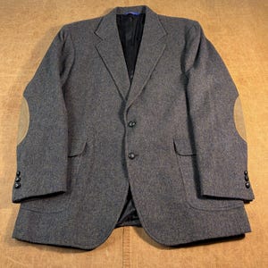 Peut inclure: Un blazer en tweed gris anthracite doté d'un revers cranté, de deux boutons sur le devant et de deux poches à rabat. Les manches sont ornées de coudières en daim beige et de trois petits boutons aux poignets. L'intérieur est doublé de noir.