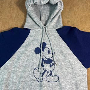 May include: Gray hooded sweatshirt with navy blue raglan sleeves. The front features a navy blue graphic of Mickey Mouse. The sweatshirt has a drawstring hood and a front pocket.