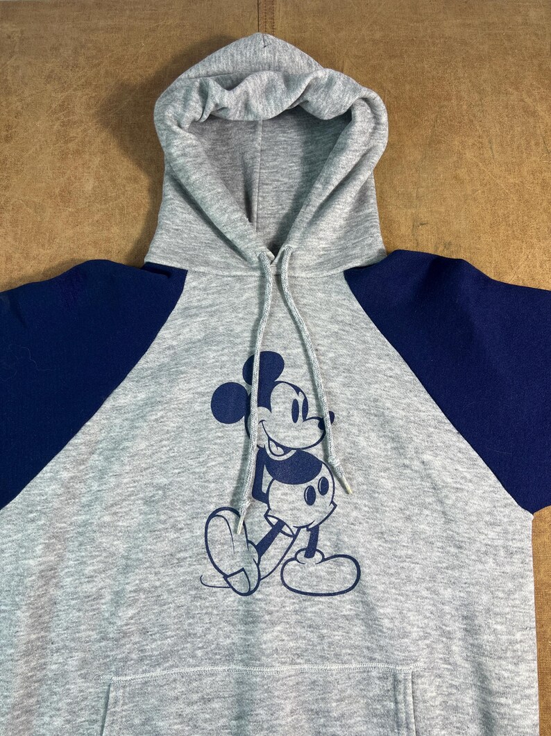 May include: Gray hooded sweatshirt with navy blue raglan sleeves. The front features a navy blue graphic of Mickey Mouse. The sweatshirt has a drawstring hood and a front pocket.