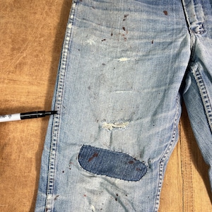 May include: Light blue denim jeans with distressed details, including a patched knee and paint splatters. The jeans show signs of wear and tear, with a faded appearance and visible stitching.
