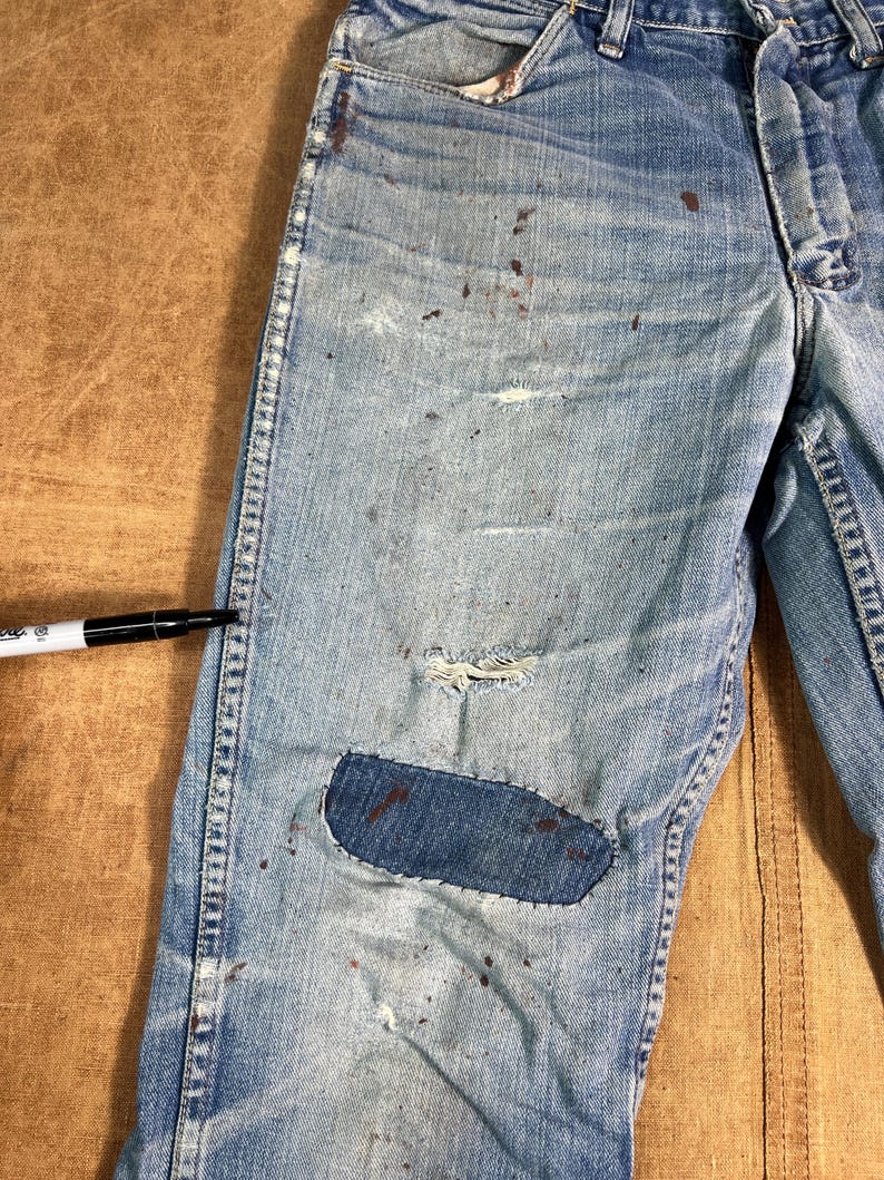 May include: Light blue denim jeans with distressed details, including a patched knee and paint splatters. The jeans show signs of wear and tear, with a faded appearance and visible stitching.