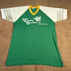 May include: Green and white vintage t-shirt with the text "Lake Country Gardens on Ottertail Lake." The shirt has a yellow V-neck collar and white sleeves with green and yellow stripes.
