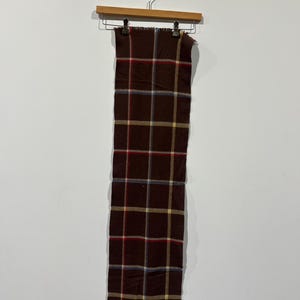 May include: A long, rectangular scarf with a brown plaid pattern, hanging from a wooden hanger. The plaid design incorporates red, yellow, and blue lines. The scarf is made of a soft material and has a fringed edge.