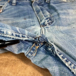 May include: Close-up of light blue denim jeans, showing the front closure and fly. The jeans have visible wear and tear, with frayed edges and distressed details. The fabric shows signs of fading and discoloration, adding to the vintage aesthetic.