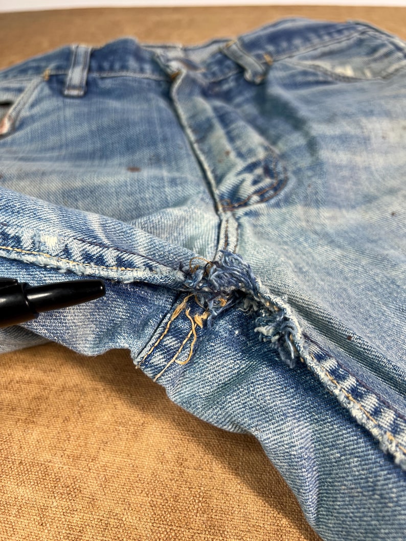 May include: Close-up of light blue denim jeans, showing the front closure and fly. The jeans have visible wear and tear, with frayed edges and distressed details. The fabric shows signs of fading and discoloration, adding to the vintage aesthetic.