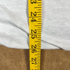 May include: A close-up shot features a yellow measuring tape with black markings against a light gray fabric. The tape shows measurements from 23 to 28 inches. The fabric appears to be a light-colored t-shirt.