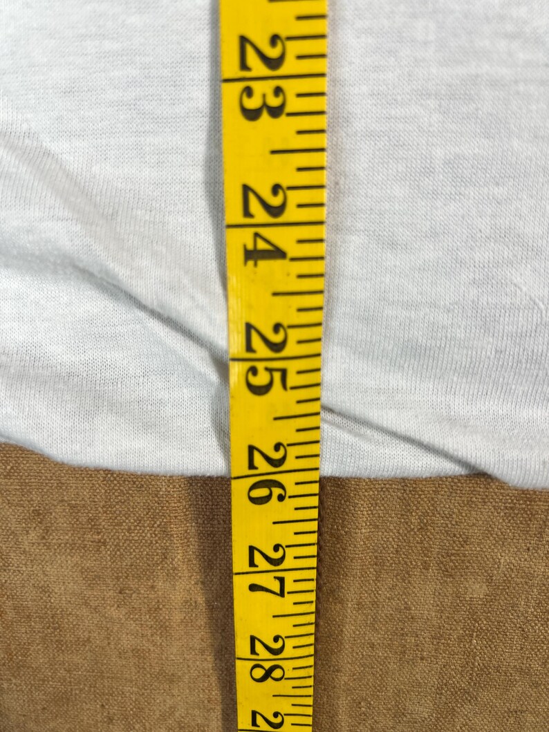 May include: A close-up shot features a yellow measuring tape with black markings against a light gray fabric. The tape shows measurements from 23 to 28 inches. The fabric appears to be a light-colored t-shirt.