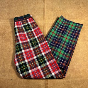 May include: A pair of plaid pants featuring a red, black, white, yellow, and blue pattern on one leg and a green, blue, and white pattern on the other. The pants are folded on a brown surface.