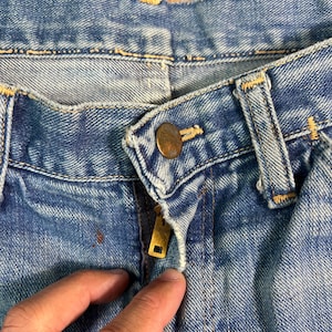 May include: Close-up of blue denim jeans with a brass zipper and button. The jeans show signs of wear and tear, with a faded appearance and visible stitching. The image focuses on the front closure and waistband.
