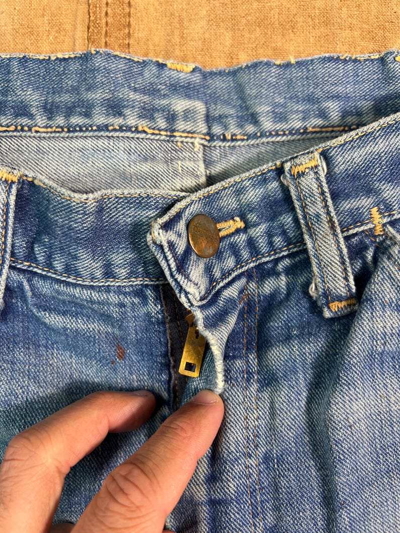 May include: Close-up of blue denim jeans with a brass zipper and button. The jeans show signs of wear and tear, with a faded appearance and visible stitching. The image focuses on the front closure and waistband.