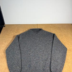 May include: A gray, speckled sweater with a ribbed collar, cuffs, and hem. The knitwear features flecks of blue and red throughout the fabric. The sweater is a classic crew neck style, suitable for cooler weather.