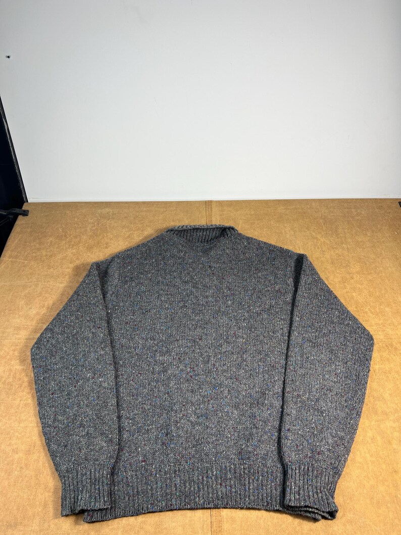 May include: A gray, speckled sweater with a ribbed collar, cuffs, and hem. The knitwear features flecks of blue and red throughout the fabric. The sweater is a classic crew neck style, suitable for cooler weather.