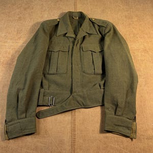 May include: An olive drab wool military-style jacket featuring a pointed collar, two large button-flap chest pockets, shoulder epaulets, and a waist belt with a metal buckle. The long sleeves have buttoned cuffs, and the garment has a structured fit.