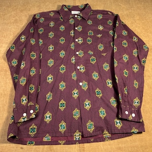 Pode incluir: Uma camisa de botão de manga comprida em um tom bordô escuro, adornada com um padrão repetido de desenhos geométricos verdes e dourados. A camisa apresenta uma gola clássica, um bolso no peito e botões brancos.
