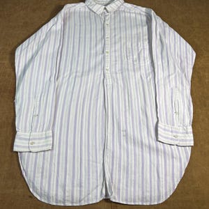 May include: White button-down shirt with vertical blue and light blue stripes. The long-sleeved shirt has a single chest pocket and buttoned cuffs. The shirt is laid flat on a brown surface.