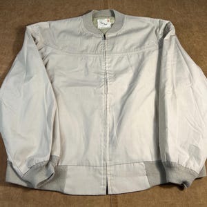 May include: A light beige bomber jacket with a full-length zipper. The jacket features a ribbed collar and cuffs in a matching light gray color. The jacket has a simple, classic design.