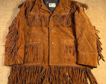 Puede incluir: Chaqueta de ante marrón con largos flecos en las mangas, el pecho y el dobladillo. La chaqueta tiene cierre de botones, dos bolsillos delanteros y un cuello. La chaqueta es de color marrón medio y parece vintage.