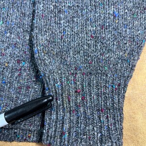 May include: Close-up of a gray knit sweater with colorful flecks. The sweater has a ribbed cuff and a visible seam. A black marker is partially visible, providing a size reference.