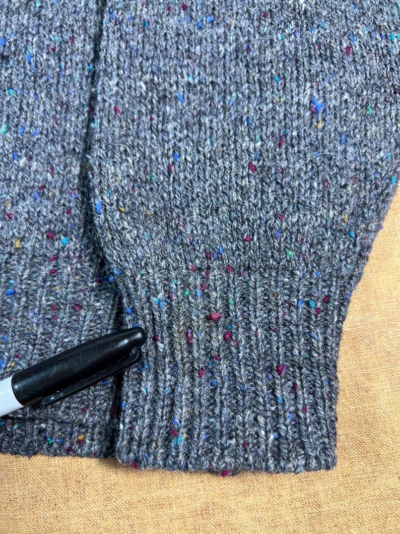 May include: Close-up of a gray knit sweater with colorful flecks. The sweater has a ribbed cuff and a visible seam. A black marker is partially visible, providing a size reference.