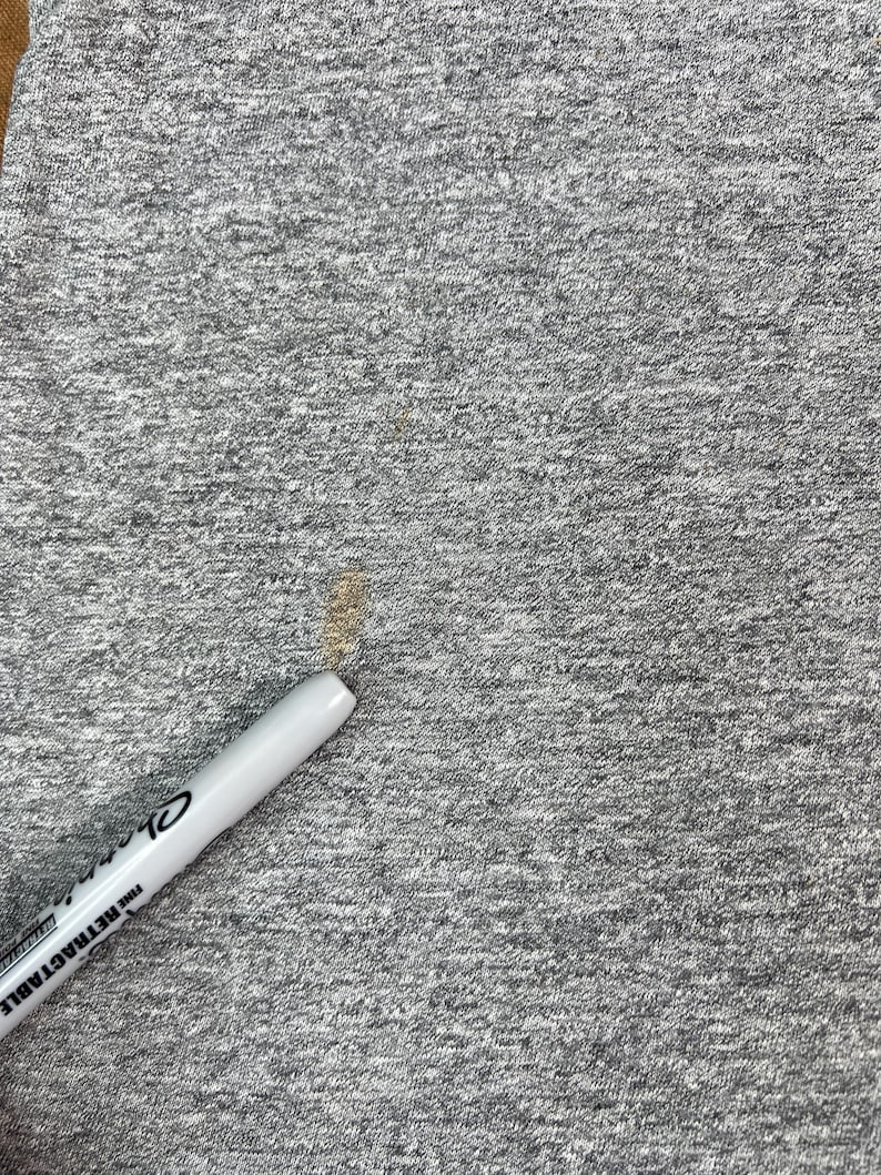 May include: Close-up of a gray, textured fabric with a white retractable pen. The pen has black text that reads "Sharpie FINE RETRACTABLE". There are brown stains on the fabric.