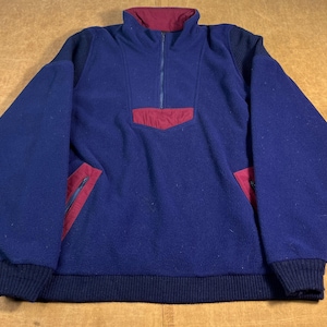 May include: A blue fleece pullover with a burgundy collar, pocket, and zipper accents. The long-sleeved top has a quarter-zip closure and two side pockets with zippers. The cuffs and hem are a darker shade of blue.