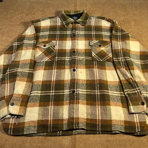 May include: A button-down, long-sleeve shirt in a plaid pattern of olive green, cream, and brown. The shirt has two chest pockets and a black collar. The cuffs are buttoned, and the shirt is made of a thick, woven fabric.