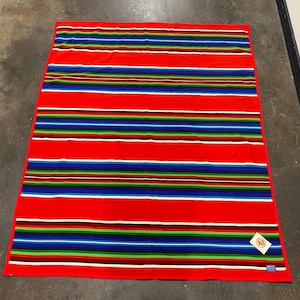 Puede incluir: Una manta rectangular roja con rayas horizontales en varios colores, incluyendo azul, verde, blanco y negro. La manta tiene una etiqueta en la esquina inferior derecha. La manta está hecha de un material suave.