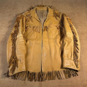 May include: A tan leather jacket featuring long fringe detailing along the sleeves, shoulders, and bottom hem. The jacket includes a pointed collar, a front button closure, and two chest pockets with flaps. The interior is lined with a smooth fabric.