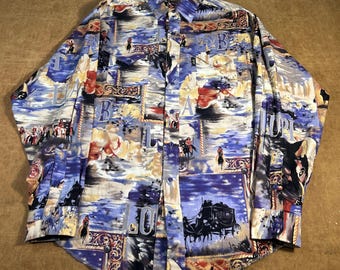 May include: Button-down shirt with a colorful Western-themed print. The shirt features a collage of images including cowboys, stagecoaches, and the letters "A", "B", and "U". The color palette includes blue, yellow, red, and black.