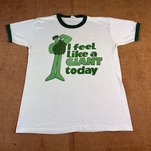 May include: A white ringer t-shirt with dark green trim on the collar and sleeves. The front features a green graphic of a character flexing its muscles, accompanied by the green text: I feel Like a GIANT today.