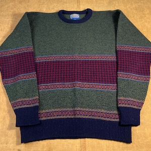 Peut inclure: Un pull vert à col rond avec un motif. Le pull présente un col, des poignets et un ourlet bleu foncé. Une large bande de motifs géométriques bordeaux et verts traverse la poitrine et les manches. Une petite étiquette est cousue sur le col.