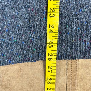May include: Close-up of a gray knit sweater with flecks of blue, red, and green. A yellow measuring tape is placed vertically along the sweater, showing measurements from 22 to 30 inches.