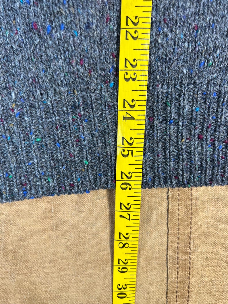 May include: Close-up of a gray knit sweater with flecks of blue, red, and green. A yellow measuring tape is placed vertically along the sweater, showing measurements from 22 to 30 inches.