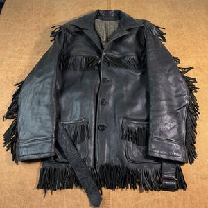 May include: A black leather jacket featuring long fringe detailing on the shoulders, chest, and hem. The jacket includes a button-front closure, two front pockets with fringe, a matching belt, and a black and white chequered interior lining.