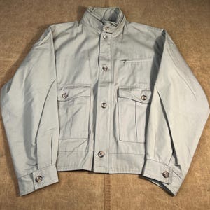 May include: A light green jacket with a high collar, button closures, and multiple pockets. The jacket has a classic design with buttoned cuffs and a small zippered pocket on the chest. The fabric appears to be a lightweight cotton or blend.