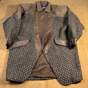 May include: A brown and black patterned coat with leather accents. The coat features a leather collar, lapels, and pocket details. The sleeves have leather cuffs with buckles. The coat has a button closure and a brown lining.