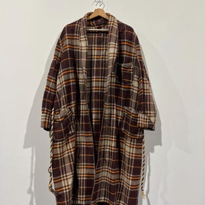May include: A long, oversized plaid robe in shades of brown, beige, and orange hangs on a wooden hanger. The robe features large pockets and rope detailing along the edges. The plaid pattern is a classic design.