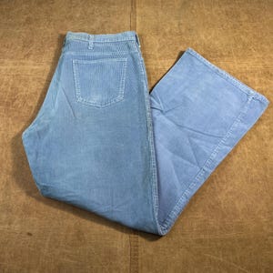 May include: Light blue corduroy pants with a classic five-pocket design. The pants are folded, showcasing the back pocket and the wide leg cut. The fabric has a textured appearance, and the color is a muted, dusty blue.