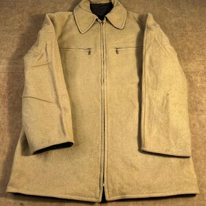 May include: A tan wool coat with a full-length zipper, two small chest pockets, and a collar. The coat has long sleeves and a black lining visible at the collar and cuffs. The coat is laid flat on a brown surface.