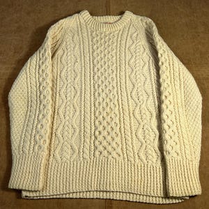 以下が含まれることがあります： クリーム色のクルーネックケーブルニットセーター。セーターは複雑なケーブル模様とリブ編みの袖口と裾が特徴です。この衣服は厚く暖かい素材でできており、寒い季節に最適です。セーターは茶色の表面に平らに置かれています。