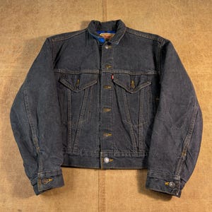 May include: A dark charcoal denim jacket featuring a pointed collar, a front button closure with metallic buttons, two chest pockets with buttoned flaps, and long sleeves with buttoned cuffs. A small red tab is visible on the left chest pocket.