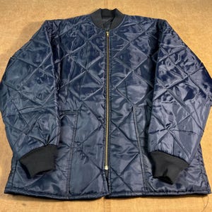 May include: A navy blue quilted jacket with a diamond pattern. The jacket has a black collar and cuffs, a gold-colored zipper, and two front pockets. The jacket is made of a shiny, smooth material.