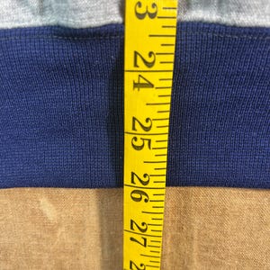 May include: Close-up of a gray and navy blue fabric with a yellow measuring tape. The tape shows measurements from 23 to 29 inches. The fabric appears to be a section of a garment.